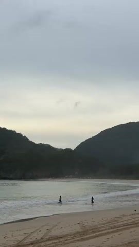 The video shows a wide shot of a beach with two people walking in the shallow water. In the background, there are green hills and a cloudy sky. The sand on the beach has tire tracks on it. One person is on the left side of the frame, closer to the water, and the other person is on the right side of the frame, further up the beach. There is a dark object on the sand to the right of the person on the right. The water has small waves rolling in.