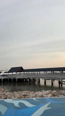 The video shows a pier with a covered walkway extending over the water. A small boat is docked next to the pier, and a larger white boat is docked further down. A small white lighthouse stands near the shore. The sky is overcast. The shore is lined with rocks, and a painted blue and white design is visible in the foreground.