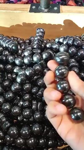 A hand holds a black, shiny, spherical object, which is one of many such objects filling a wooden bin. The objects are magnetic and appear to be hematite stones or magnetic marbles. The hand manipulates the object, showing it to the camera before dropping it back into the bin. The bin is filled with hundreds of these objects, creating a large pile. In the background, other colorful stones or marbles are visible in a separate section of the bin. The lighting suggests an indoor setting, possibly a store or market.