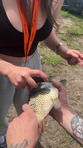 Here is a description of the video.

The video shows a person holding a fish while another person cleans it. The fish has a patch of blue paint or dye on its scales. The person cleaning the fish uses a small tool to remove debris from inside the fish’s mouth. The person holding the fish has tattooed arms. The cleaner is wearing a black top, gray sweatpants, a watch, and a lanyard with “FORREST” printed on it. The video takes place outside on a grassy area.
