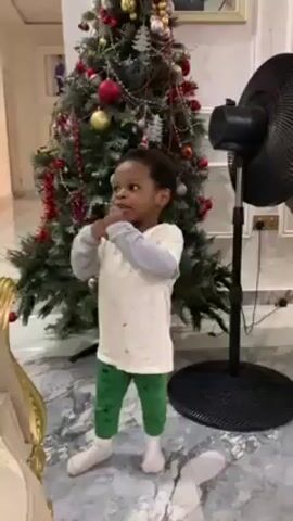 A young child stands in front of a Christmas tree in a house. The child wears a white long-sleeved shirt with a floral pattern, green leggings, and white socks. The Christmas tree is decorated with red and gold ornaments, tinsel, and lights. A black standing fan is visible near the Christmas tree. The child smiles and claps, then climbs onto a sofa. The child then looks up at the wall. The floor is light marble.