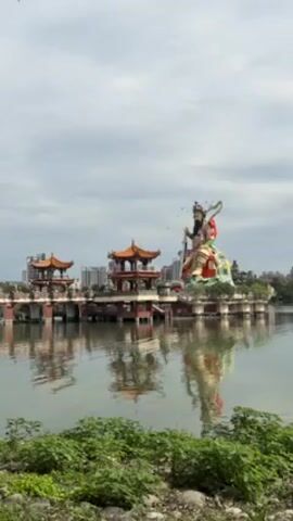 The video shows a view of Lotus Pond in Kaohsiung, Taiwan. The body of water reflects the buildings in the background.  Two traditional Chinese style pagodas connected by a bridge are visible on the left side.  On the right is a large, colorful statue of a deity sitting on what appears to be a cloud or lotus-like base.  The sky is overcast, and the foreground features green foliage.