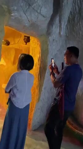 The video shows two people inside a cave-like structure with textured walls. A woman in a white shirt and blue skirt stands facing a brightly lit yellow alcove containing various objects, some of which appear to be masks or figurines. A man in a blue shirt stands to her right, holding up a smartphone and appearing to take a picture. The cave walls have a rough, uneven texture, and there are some stalactite-like formations hanging from the ceiling. The lighting is dim in most of the cave, with the yellow alcove being the primary light source.