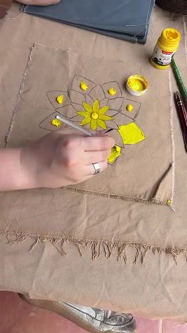 A close-up, overhead shot shows a person's hands painting a flower design on a piece of light brown fabric. The fabric has visible texture and frayed edges. A dark outline of a multi-petaled flower or mandala is drawn on the fabric. The person uses a small paintbrush to apply bright yellow paint, scooping it from a small white container with a yellow lid. Several petals of the flower have already been filled with the yellow paint, and the person is actively painting additional petals, carefully staying within the lines of the drawn outline. The paint is applied with some thickness, creating texture on the fabric. A partially visible yellow bottle of paint in the background has the word "ACRYLIC" printed on it. A dark gray piece of fabric and a pen or pencil are also visible in the background. The person wears a silver ring on their left hand. The painting appears to be in progress.