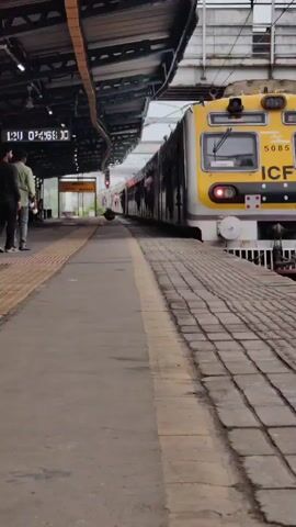 A train is stopped at a station. The train is yellow and white. There are people on the platform and in the train cars. The train is under a covered platform. There is a sign that says "DAHISAR". The camera is at a low angle, looking up at the train.