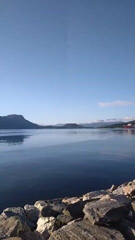 This is a vertical video of a calm body of water, likely a fjord or a large lake, with mountains in the background. The water is dark blue and reflects the clear, pale blue sky. In the foreground, there is a rocky shoreline. In the water, there are a few objects: a yellow buoy on the left, and a small, dark inflatable boat with a white console in the center. Further in the distance, on the left side of the frame, a wooden pier or jetty is visible with some posts. The sun is shining brightly, creating a glare in the upper left portion of the sky. The scene appears peaceful and serene.
