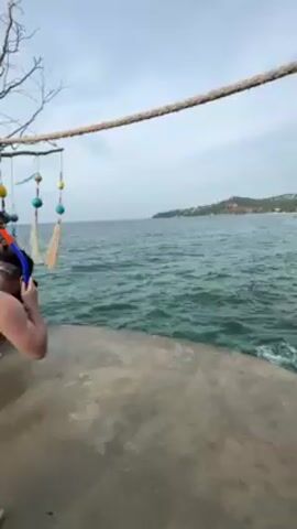 The video begins with a mid-shot of a fair-skinned man in khaki shorts and snorkeling gear jumping into a body of water from a concrete platform. As the man leaps, the camera pans to a decorative structure crafted from tree branches adorned with painted wooden paddles and colorful orbs.  A person with long dark hair and a green hat briefly covers their face with their hand as they are filmed from below. The scene shifts back to the water, where bubbles indicate where the man landed. In the background, there is a lush green coastline dotted with buildings, silhouetted against a cloudy sky. The water is a vivid shade of turquoise, with gentle waves lapping against the platform's edge.