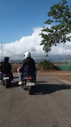 The video shows two people riding a motorcycle on a dirt road. The person in the back is wearing a white helmet and a dark jacket. The motorcycle is black and has a license plate that reads "AB 4008 CI". The road is lined with dry grass and a fence with barbed wire. There are some trees in the background, including a palm tree. The sky is blue with some clouds. The video appears to have been taken during the day.