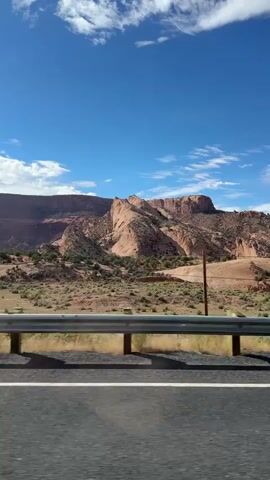 A side-view video captures a landscape from a moving vehicle. The foreground shows a segment of a paved road with a white line marking the edge, followed by a silver metal guardrail. Beyond the guardrail, a dry, brush-covered terrain slopes upward towards a majestic, arid mountain range. The mountains are characterized by rugged, eroded rock formations, primarily in shades of red and tan, with sparse vegetation clinging to their slopes. The sky above is a vibrant blue, dotted with scattered white clouds, indicating a clear, sunny day. The vehicle appears to be traveling along a highway that runs parallel to the mountains. There are no people or distinct actions visible, only the continuous, static view of the natural environment passing by.