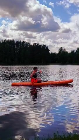 A person is sitting on an orange paddleboard in the middle of a lake. The person is wearing a life vest and orange shorts, and they are holding a paddle. The paddleboard has some writing on it. The background shows a line of trees and a sky with clouds. The water is rippling, and reflections of the sky and trees are visible. The person is paddling.