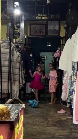 Two young girls are in what appears to be a clothing store. The girl on the left wears a pink tutu and leotard, while the girl on the right wears a pink pajama set. They are interacting with a blue child's toy, possibly a ride-on toy or a push toy with a handle, in the middle of an aisle. The store has racks of clothing, wooden shelves, and ornate lanterns. The floor is made of dark, worn wood. The overall lighting is dim, suggesting it might be late in the day.