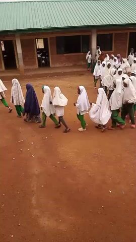 A group of children, mostly wearing white hijabs and green pants, are walking in a line in an outdoor area. The ground is dirt, and there is a building with a green roof and brick walls in the background. Some of the children are holding objects in their hands. The line of children is moving from left to right in the frame. Another group of children is standing near the building.
