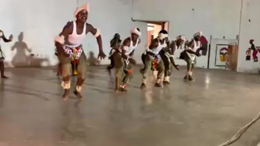 A group of about eight barefoot male dancers is performing a rhythmic dance inside a white-walled room with a smooth concrete floor. They are clad in white tank tops, olive-green trousers, and decorated skirts or aprons, and sport white headbands. Most dancers are aligned in a row, rhythmically bending forward and back in unison, while one dancer, dressed similarly, performs individual steps at the left of the screen. In the background, there are abstract drawings on the wall, a doorway, and a small pile of coiled rope on the floor. The general light is diffused but bright.
