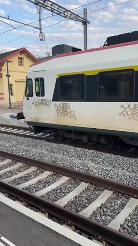 A train car with graffiti on its side is visible at a train station. The train car is white with a yellow stripe and black windows. The graffiti is in a light color, possibly white or light grey, and appears to be in a stylized font. The train is on a track with gravel ballast. In the background, there is a building with a red roof and a light-colored facade. Overhead, there are electrical wires and poles for the train. The sky is cloudy. The camera is positioned at eye level, slightly angled down towards the train.