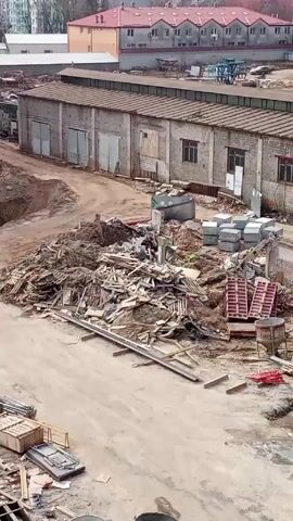 The video shows a construction site with piles of lumber and debris in the foreground. In the background are low brick buildings with multiple garage doors, and taller residential buildings. A person walks along a dirt path towards the brick buildings. There is some machinery or equipment visible in the distance beyond the brick buildings. The ground is mostly dirt and gravel. There is a partially constructed building to the left of the frame. The video is taken from an elevated position, possibly from another building.  The scene appears to be an area under redevelopment or demolition and construction.