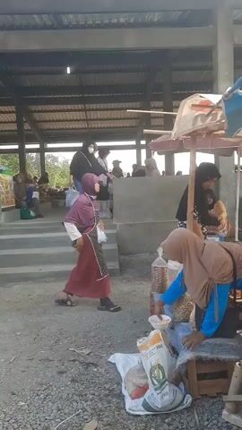 A street market is depicted. In the foreground, a woman wearing a brown headscarf and blue shirt bends over a market stall. She is reaching into a plastic bag of nuts. In front of the stall, there is a basket filled with bottles of juice. Behind the stall, a woman in a black headscarf and floral dress stands and observes. Further back, several people are visible in the open-air market, with some standing on elevated steps and others sitting. The market appears to be covered by a corrugated metal roof supported by wooden beams. The ground is covered with gravel and dirt.