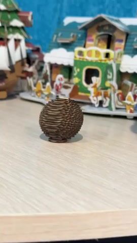 A short close-up video shows a cardboard Christmas village scene. In the foreground, there is a brown, spherical object, composed of layered cardboard. Next to it, a cardboard sculpture of a bulldog is placed on a light-colored surface, possibly a shelf or table. In the blurred background, a colorful Christmas village building is partially visible. The background is a solid blue color.