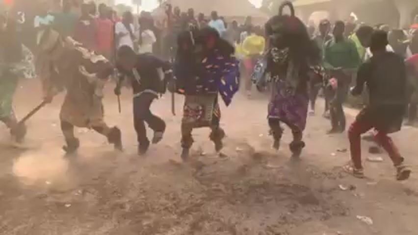 The video shows a group of people in what appears to be a village or rural area. Several individuals are wearing elaborate, colorful costumes and masks that obscure their faces. These costumed figures are actively moving and dancing, kicking up dust from the ground. Some appear to be holding sticks or rods. A crowd of onlookers surrounds them, observing the performance. The environment is dusty and appears to be outdoors, with some buildings or structures visible in the background. The overall atmosphere seems celebratory or ceremonial.