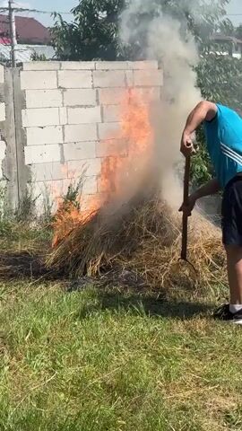 A man in a blue t-shirt and dark shorts is standing to the right of a large pile of dry hay that is actively burning. He is holding a long-handled tool, possibly a rake or a hoe, and appears to be tending to the fire. The fire is producing a significant amount of white smoke and bright orange flames that reach upwards. Behind the fire and the man is a wall constructed of white concrete blocks. To the left of the concrete wall, a house roof is visible. In the background, there are green trees and a clear blue sky. The fire is burning on a patch of green grass.