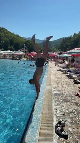 A man is captured mid-air performing a somersault dive into a swimming pool. The camera pans to follow his descent, showing the splash as he hits the water. The pool is surrounded by lounge chairs and umbrellas, with a water slide visible in the background. Lush green hills and a clear blue sky complete the scenic backdrop.