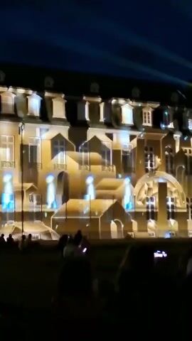 The video shows a nighttime outdoor scene with a crowd of people silhouetted in the foreground, facing a large building illuminated by a light projection show. The building appears to be a historical structure, possibly a palace or grand house, with numerous windows and architectural details. The projection transforms the facade into an animated display, featuring geometric shapes, faux windows, and streaks of light, creating an illusion of depth and movement. Bright beams of light emanate from the sky and project onto the building, adding to the dynamic visual effect. The crowd is diverse, with individuals holding up phones to record the spectacle. The overall atmosphere is one of awe and entertainment.