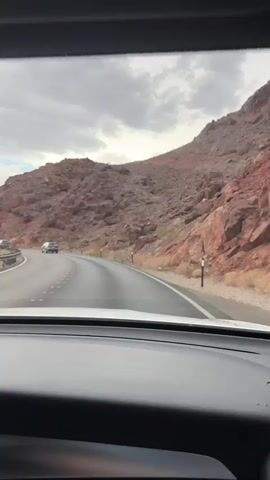 The video shows a scenic drive through a mountainous, desert-like landscape, likely near Boulder City, United States. The viewpoint is from inside a car, looking out through the windshield. The road curves to the left, with a steep, rocky, reddish-brown mountain face dominating the right side of the frame. The left side of the road features a metal guardrail and a precipitous drop-off, with a vast, layered mountain range visible in the distance under a cloudy sky.

Several cars are visible on the road ahead, indicating ongoing traffic. The sky is overcast with dramatic clouds, suggesting potentially changeable weather or a late afternoon/early morning light. The overall mood conveyed is one of exploration and enjoying a scenic route, with the vastness of the landscape inspiring a sense of awe.

A significant detail is a digital display on the car's dashboard on the lower left of the screen. It shows "35 33" on the left and "NW" on the right. This likely indicates the vehicle's internal temperature and compass direction. There are also navigation prompts visible on the screen: "In half a mile, turn left to stay on Nevada 172." This confirms the location is indeed Nevada and provides a specific route detail. The presence of power lines and poles along the road also points to infrastructure in a somewhat remote area. The sun is not directly visible, but the diffuse light suggests it is not midday.