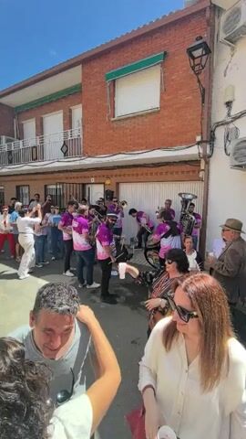 This video captures a vibrant daytime street celebration in Fontanarejo, Spain. In the immediate foreground, a couple is seen sharing a kiss, their faces close together. Behind them, the main focus is a lively brass band performing in the middle of a narrow residential street.

The band consists of approximately 10-15 musicians, all wearing matching purple short-sleeved shirts with white designs, playing a variety of brass instruments including trumpets, trombones, and a tuba. They appear to be actively engaged in playing music, contributing to a festive atmosphere.

A crowd of people of various ages is gathered around the band, listening to the music and socializing. Many individuals are holding plastic cups, and some are seen dancing or moving to the rhythm. The mood is clearly joyous and celebratory.

The street itself is paved and lined with two-story houses, primarily constructed of red brick. One house in particular features white-railed balconies and closed green window shutters or awnings. The sky is bright blue, and the strong sunlight indicates a clear, sunny day, likely midday or early afternoon. The scene evokes a sense of community, music, and celebration. No specific text is clearly legible in the video.