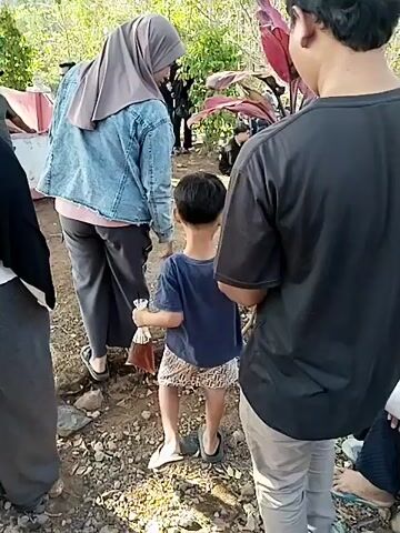 The video shows a group of people gathered outdoors, likely in a wooded or natural area. The ground is covered with dirt, rocks, and some sparse vegetation. Several individuals are seated on the ground, some wearing hijabs. In the foreground, a young child in a blue shirt and patterned shorts is being led by an adult. Behind the child, another adult in a black t-shirt is visible. Further in the background, more people are seated, and a few are standing. There is a pink umbrella visible in the background. The overall atmosphere appears to be a casual gathering or event.