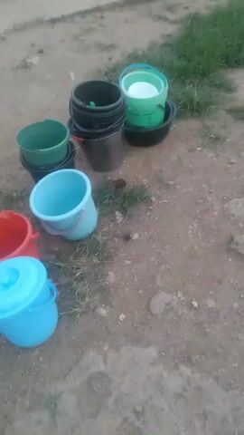 The video shows a collection of variously colored plastic buckets and tubs scattered on a dirt and gravel surface. The buckets and tubs are of different sizes and stacked in some places. A person wearing a denim skirt and sandals walks behind the collection in the background. The environment appears to be an outdoor area, possibly a yard or a clearing near a building. Some sparse patches of green grass are visible.