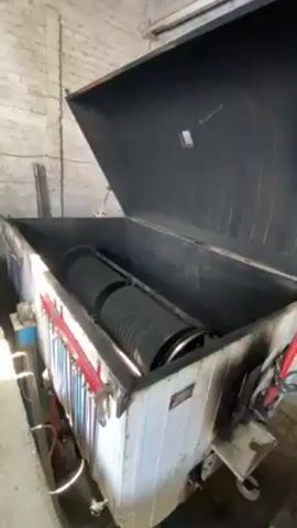 The video shows a large, box-shaped machine with its lid open in what appears to be a warehouse or industrial setting. Inside, a cylindrical object with ribs or ridges is visible, presumably part of the machine's mechanism. The machine is white with some rust and wear, and various pipes and controls are attached to its side. A man in a brown outfit walks into the frame in the background, moving from left to right. The warehouse has white brick walls.