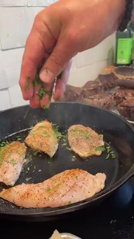 A person wearing a white chef's jacket with red trim is in a kitchen. They are in the process of cooking, as evidenced by the cast iron skillet with chicken breasts on the stovetop. The person is sprinkling chopped herbs onto the chicken. In the background, several steaks are visible on a cooling rack, along with bottles of sauces and other kitchen essentials. The kitchen appears to be well-lit and modern, with white tiled walls and cabinets.