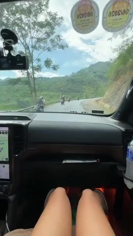 The interior of a car is captured in this video with the driver's perspective. The car's dashboard is prominently visible, and a GPS screen displays a route. In front of the dashboard, a pair of legs is visible. A water bottle sits in a holder on the side. Stickers are affixed to the windshield. Outside the car, the road winds through a mountainous area with lush green vegetation. Motorcyclists can be seen driving on the road ahead. The sky is partly cloudy.