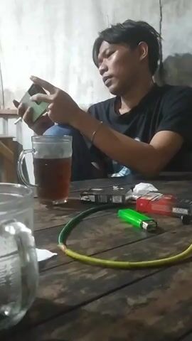A man with short black hair and a black t-shirt sits at a wooden table, looking down at a mobile phone in his hands. He is holding the phone with both hands, with his thumbs positioned on the screen. To his left is a clear mug filled with a dark liquid, likely tea. On the table, in front of him, are two packs of cigarettes, one red and one green, and a bright green lighter. A coiled green and yellow hose lies on the table as well, possibly part of a hookah setup. Another clear mug, this one empty and with visible markings, is partially in the foreground on the left side of the frame. The background appears to be a rough, textured wall. The lighting is dim, suggesting an indoor setting with limited illumination.