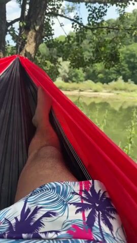 The video begins with a close-up shot of a person's legs from the knee down, wearing patterned shorts, as they lie in a red and black hammock. The camera then pans out to reveal the view from the hammock, which overlooks a tranquil body of water surrounded by lush green trees and vegetation. The sun is shining, casting a soft light on the scene, and the sky is partially visible through the canopy of leaves. The camera then shifts focus slightly, showing more of the person's legs and feet, as well as the details of the hammock itself. The video continues with a slow pan across the landscape, capturing the serene beauty of the natural environment. The water reflects the surrounding greenery, and the gentle sway of the hammock suggests a peaceful and relaxing atmosphere. The video ends with a final shot of the person's legs and feet, still resting in the hammock, as the camera pulls back to encompass the entire view.