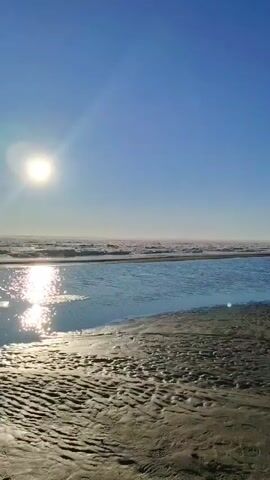 This is a vertical video of a sunny beach at low tide. The sun is shining brightly in the clear blue sky, casting a reflection on the shallow water. The sand is wet and textured, with ripples from the receding tide. In the distance, the ocean waves are visible, and a hazy horizon suggests the presence of more water or land. There are no people or objects visible in the scene.