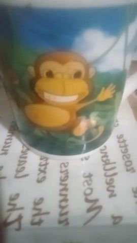 The video displays close-ups of a mug with a monkey illustration and other objects on a patterned surface. The mug features a cartoon monkey with a yellow face, large brown eyes, and a wide smile, clinging to a green vine against a blue and green background. In other frames, a dark liquid, likely coffee or tea, fills a white mug, and there are glimpses of other mugs and what appear to be fruit slices and a bottle on the patterned surface. The overall scene suggests a casual setting, possibly a kitchen or dining area, with a slightly blurry, handheld camera perspective. No specific time of day or weather conditions are discernible. The visible text on the surface is in a font that appears to be reversed or reflected, making it difficult to read, but some words like "rosette," "wellness," "most," and "extra" might be partially discernible. There are no people visible in the video, and the focus is on the objects and the mug's illustration. The emotions conveyed are neutral, centered on the depiction of the playful monkey on the mug.