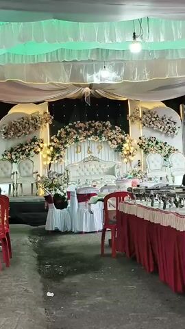 A video recording of an outdoor wedding reception stage. The stage is decorated with white and pink flowers, and has a white sofa and two white chairs. In front of the stage, there are tables with red tablecloths and food. The ground is paved with a grey material. There are some people present, but they are mostly out of focus or in the background. The overall lighting is dim, with some colored lights on the ceiling.