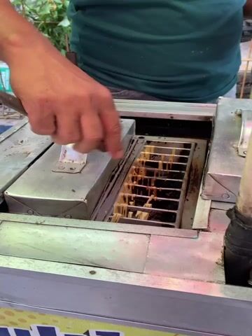 A person's hand is using a metal tool to separate items that are cooking in a metal apparatus. The apparatus appears to be a commercial food preparation device, possibly for making street food. The items being cooked are light brown and appear to be in individual slots within the apparatus. The hand is wearing a ring on its ring finger. The person is wearing a green shirt. The environment seems to be outdoors, with greenery visible in the background and what appears to be a cart or stand that the apparatus is mounted on.