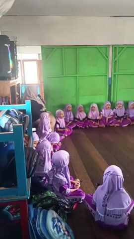 A group of young children, dressed in matching purple hijabs and outfits, are sitting in a circle on a wooden floor. They appear to be in a classroom or play area, with green walls visible in the background. Several adults, also wearing hijabs, are present, supervising the children. Some of the children are looking towards the adults, while others are interacting with each other. There are some bags and what appear to be cups stacked on a table in the background. The overall atmosphere seems calm and engaging, suggesting a learning or activity session.