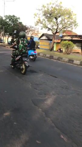 Two people are riding scooters on a paved road. The scooter in the foreground is black with gold accents and is being ridden by a person wearing a camouflage uniform and a green helmet. The scooter in the background is blue and is being ridden by a person wearing a dark blue jacket and a dark helmet. The road appears to be in disrepair with visible cracks and patches. Trees and buildings line the side of the road. The time of day appears to be late afternoon or early evening due to the lighting.