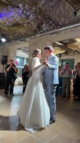 A wedding reception is captured in this video. The bride, in a white wedding dress, and the groom, in a light blue suit, are dancing in the center of the frame. They are surrounded by a bubble machine, creating a whimsical atmosphere. Several guests are visible in the background, some holding phones to record the moment. The venue appears to be an indoor space with a high ceiling and exposed beams. The lighting is subdued, with spotlights highlighting the dancing couple.