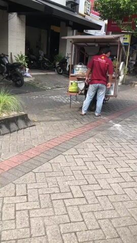 The video shows a man standing by a food cart on a paved walkway. He is wearing a red shirt with yellow lettering and blue jeans. The food cart is a mobile unit with a canopy and shelves, containing what appear to be steamers. A green gas cylinder is attached to the side of the cart.

The scene appears to be an outdoor street environment. The walkway is made of interlocking pavers with a red and gray pattern. In the background, there are buildings, some with visible signage. One sign reads "WAKAM". There are also plants in pots and what looks like motorcycles parked nearby.

The man is facing away from the camera, looking towards the food cart, and seems to be attending to it. The activity suggests a vendor preparing or serving food from a mobile stall.

The lighting indicates it is daytime, likely morning or afternoon. The weather appears clear and sunny, given the shadows. The location cues, such as the architecture and the style of the food cart, are consistent with a street scene in Southeast Asia, specifically East Java as indicated. There are no explicit emotional cues visible from the man's posture.

The visible text on the man's shirt appears to be "Sari Rasa".
