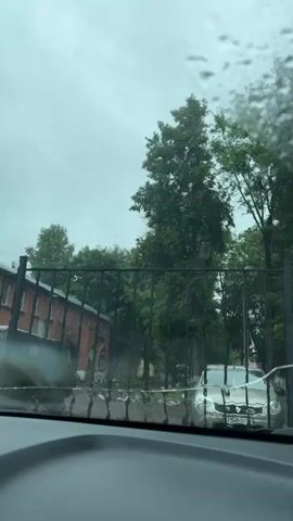 The video shows a view through a rain-streaked car windshield. In the foreground, the dashboard of the car is visible. Through the windshield, a black metal fence is prominent. Beyond the fence, a brick building with arched windows is visible on the left. Green trees form a dense backdrop. A light-colored car is parked on the right side of the scene. The sky is overcast and gray, indicating rainy weather. The trees appear to be swaying, suggesting wind. The overall atmosphere is subdued due to the weather conditions.