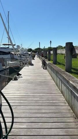 A person walks down a wooden pier lined with boats on the left and a grassy area with some chairs on the right. The sky is clear and blue. A white sailboat is docked on the left, with other boats visible behind it. The pier has a wooden railing. In the distance, there are trees and buildings. The person is wearing dark clothing.