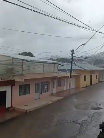 The video shows a residential street during heavy rainfall. The scene is viewed from an elevated position, possibly a window. The street is wet and there are puddles forming. Several single-story houses line the street, painted in light colors like peach and yellow. Water is seen gushing from a pipe near a utility pole, adding to the street's already drenched state. Overhead, a dense network of power lines stretches across the frame against a grey, overcast sky. The distant hills are barely visible through the rain and low clouds.
