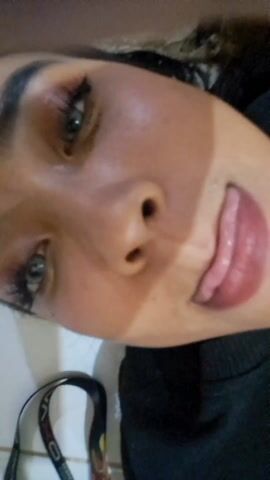 The video shows a close-up of a woman's face. She has dark hair and light-colored eyes. Her skin appears to have a tan line or shadow on it. She is wearing makeup, including eyeshadow and lipstick. The background is blurry and indistinct.