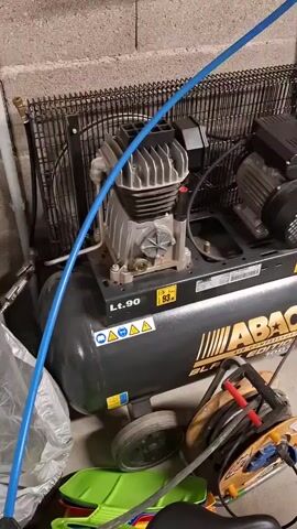 Here is a description of the video:
The video shows a black and gray ABAC air compressor with a blue air hose and a blue retractable hose reel attached to a cement block wall. The air compressor is labeled "Lt.90" and "HP 2." It is sitting on a concrete floor in a room with a cinderblock wall. Some other objects are also near the air compressor, including a green sled, a cable reel, and a gray pipe. The air compressor is running.