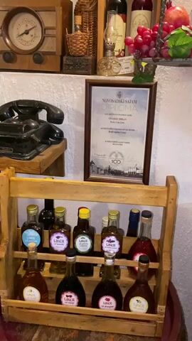 The video pans across a rustic display of various artisanal products and vintage decor, likely within a shop or a tasting room in Meterizi, Montenegro.

The scene is characterized by wooden shelving and furniture, set against a white textured wall. On the top shelf, an antique wooden radio with a large circular dial and an old-fashioned oil lamp are visible. Below, an antique black rotary telephone rests on a wooden shelf.

Framed diplomas with red ribbons, bearing the text "NOVO SAD SAJAM DIPLOMA," are prominently displayed on the wall, suggesting awards or recognition for the products.

In the foreground, a multi-tiered wooden rack holds numerous small bottles labeled "LIKER." These bottles feature different colored labels and appear to represent various flavors, such as "LAVANDER" and "RUŽA." Other bottles of varying sizes, including larger ones of wine or spirits, and small jars of preserves are arranged on and around the wooden furniture. A small wooden barrel is partially visible at the bottom.

Other decorative items include a silver metal pitcher, a candelabra, and an arrangement of grapes and a pomegranate. The overall ambiance is traditional and emphasizes local, handcrafted goods.
