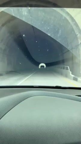 A car is driving through a tunnel. We see the view from inside the car, looking out of the windshield. A car is ahead. The tunnel is concrete and dimly lit, with lights overhead.