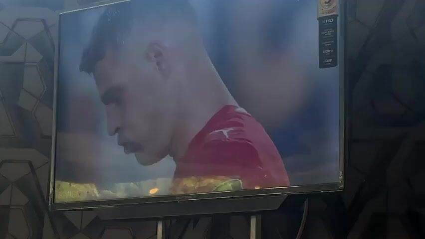 A television screen displays a soccer player in a red jersey, head bowed and on one knee, with his right fist clenched. The player appears to be reacting to a goal or a missed opportunity. The screen is mounted on a wall covered in a geometric patterned wallpaper. The lighting is dim, with reflections visible on the screen.